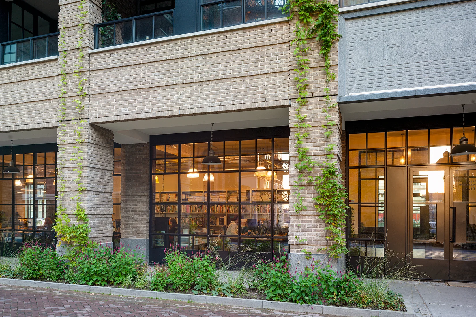Gevel van een modern gebouw aan de Karl Weisbardstraat met grote ramen, klimplanten en zicht op een bibliotheekachtige ruimte binnenin.